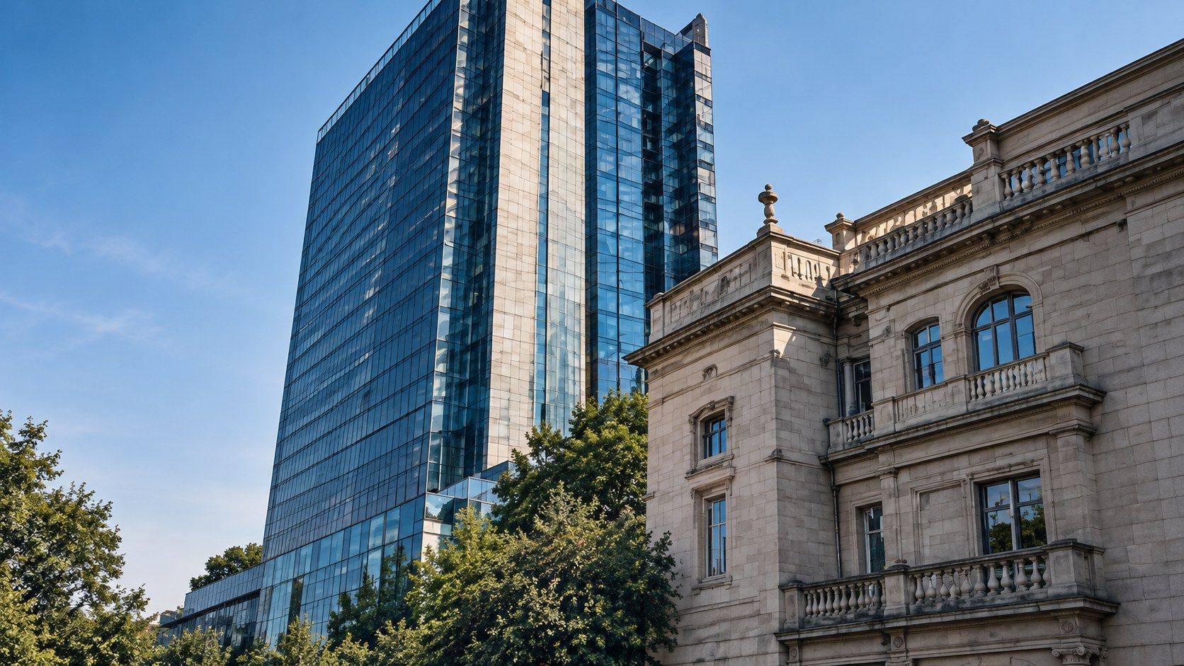 A modern glass tower rising behind a classical stone building with balustraded balconies, framed by trees against a clear blue sky